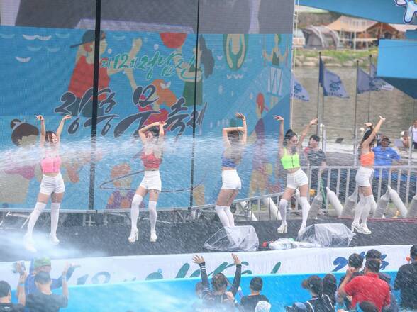 지상최대의 물싸움장 무대에서 여성 5명이 춤을 추고 있고 하단의 풀장에서는 관람객들이 물을 맞으면 뛰놀고 있다.
